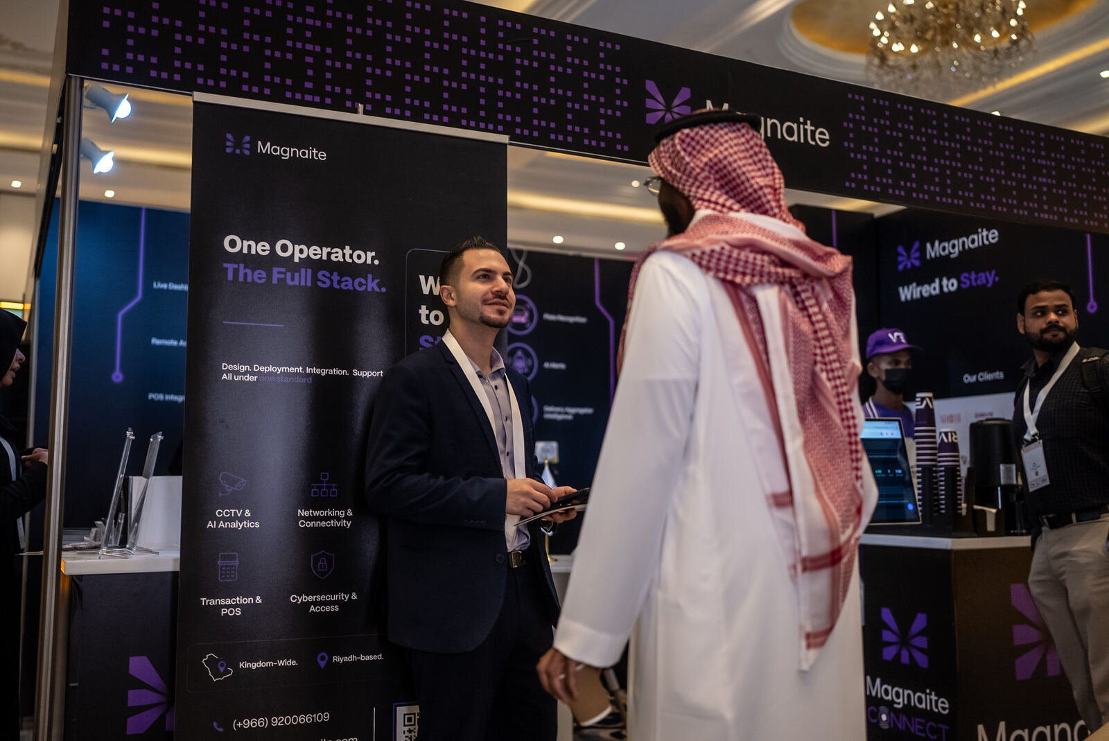 A Magnaite team member speaking with a visitor at the booth, 'One Operator. The Full Stack.' signage visible.