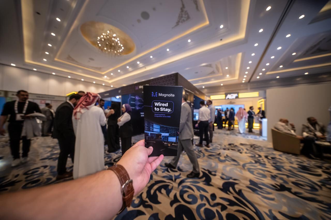 Holding up the Magnaite 'Wired to Stay' brochure on the event floor, booth visible in the background.